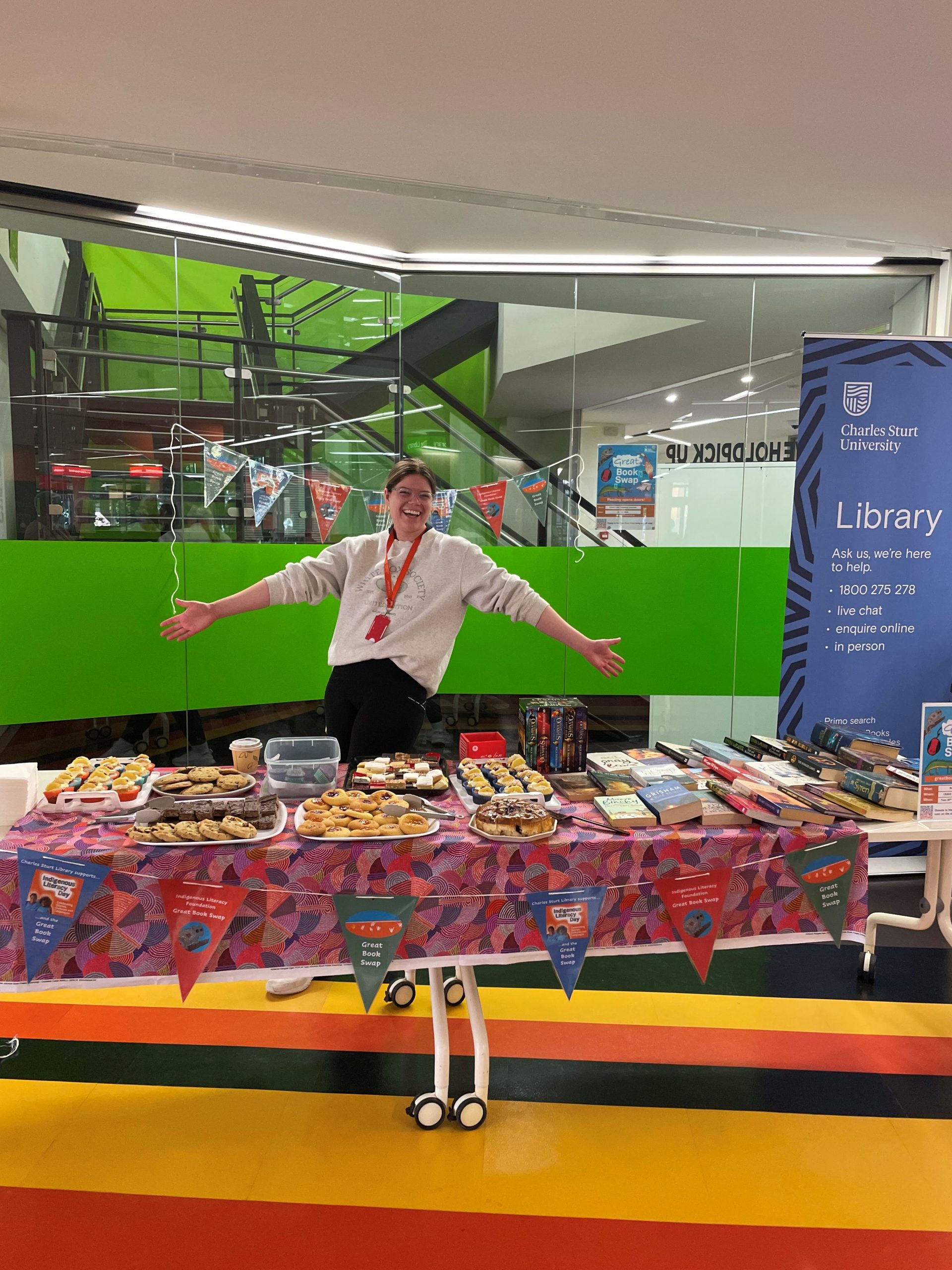 Library staff members at the Great Book Swap staff in Wagga.  She is standing with her arms out behind a table covered in cakes and biscuits.