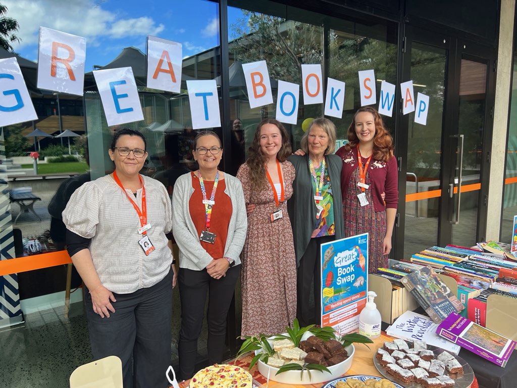Library staff members Tracie, Lorraine, Natasha, Alison and Jessica at the Great Book Swap stall in Port Macquarie. They are standing behind a large table filled with books and cakes.