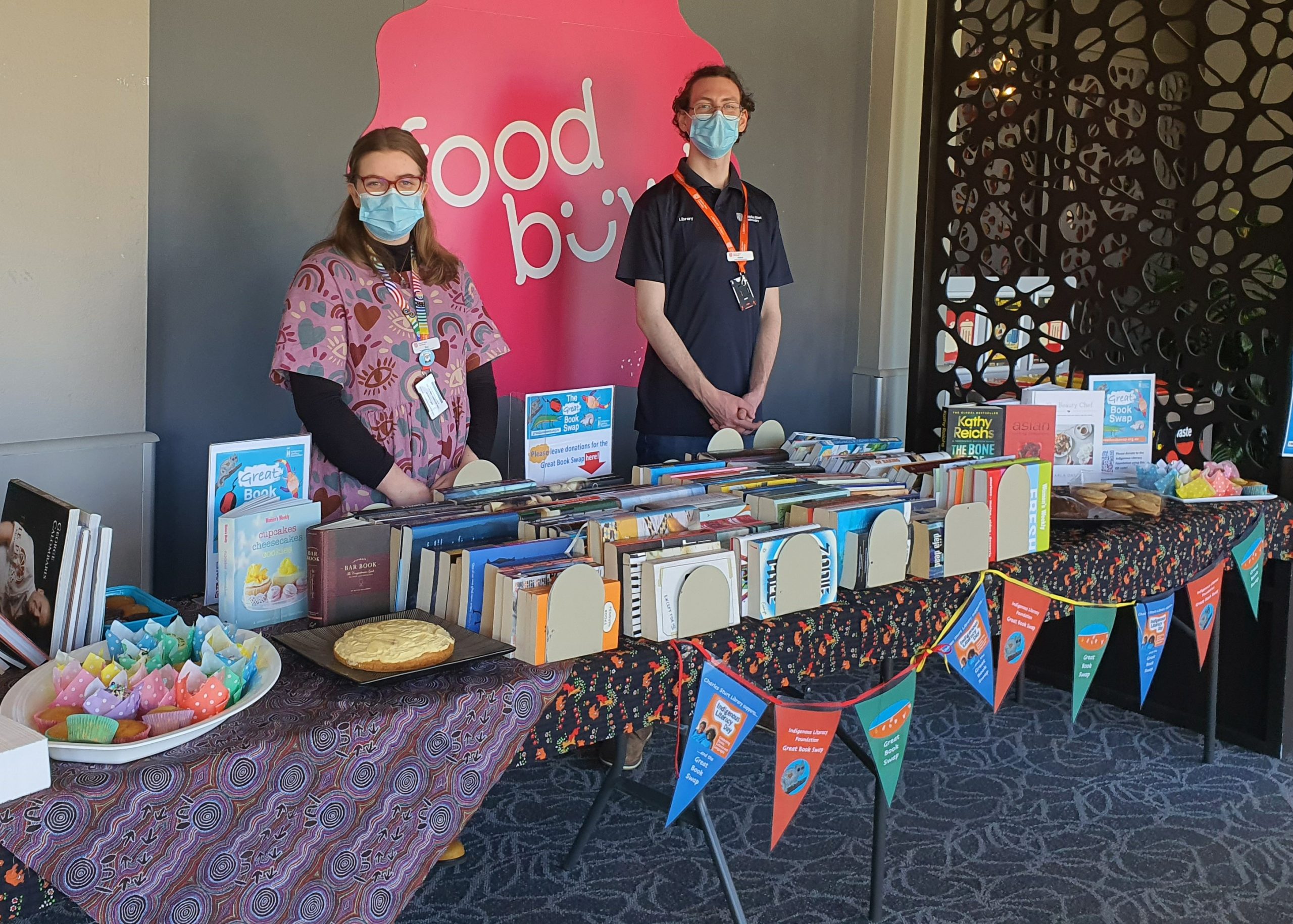 Library staff members Bea and Pat at the Great Book Swap stall in Bathurst. They are standing behind a large tables filled with books and cakes.
