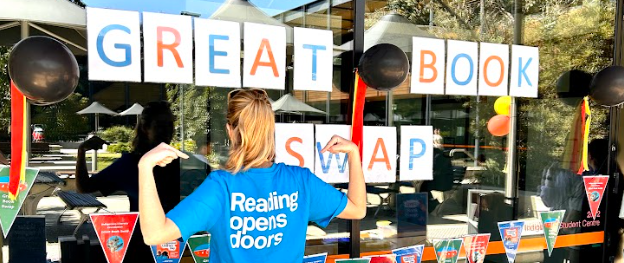 Woman with her back to the camera, pointing at the slogan on the back of her t-shirt; "Reading Opens Doors".